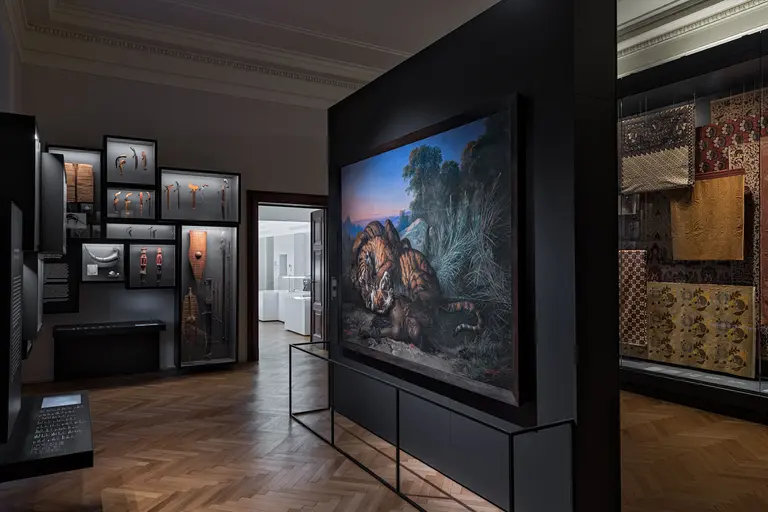 Das Bild zeigt einen Innenraum eines Museums mit einer großen Darstellung eines Löwen und üppiger Vegetation. Umgeben von Vitrinen mit Exponaten, vermittelt es eine kulturelle und historische Atmosphäre. Der Boden ist aus Holz, und die Wände sind dunkel gehalten, um die Kunstwerke hervorzuheben.