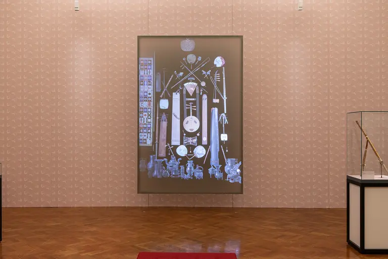 The image features a large illuminated display in a gallery, showcasing various historical artifacts and symbols. Surrounding the display, the floor is made of polished wood, enhancing the exhibition's formal atmosphere. A glass case nearby contains additional items of interest related to the artifacts on the wall.