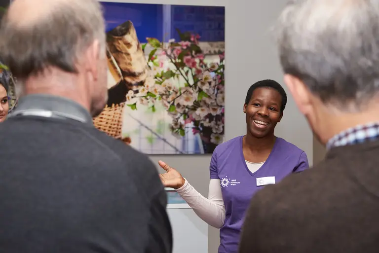 Eine lächelnde Frau in einem lila Shirt spricht mit einer Gruppe von Menschen. Sie steht in einem hellen Raum mit einem bunten Hintergrund, der Blumen zeigt. Die Zuhörer scheinen interessiert zuzuhören.