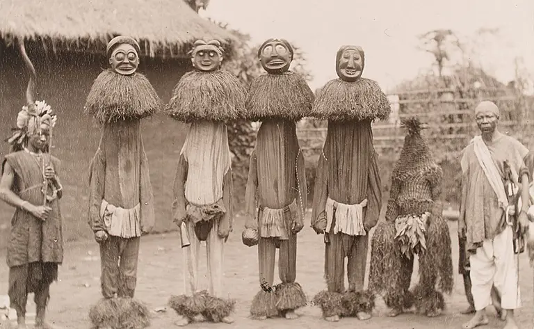 Sechs Personen stehen in einer Reihe, tragen traditionelle Kostüme und Masken. Einige haben lange Kleidung aus Pflanzenmaterial, andere tragen spezielle Kopfmasken. Im Hintergrund sind eine Birkenhütte und eine Landschaft zu erkennen. Ein Mann auf der linken Seite hält einen Stock.