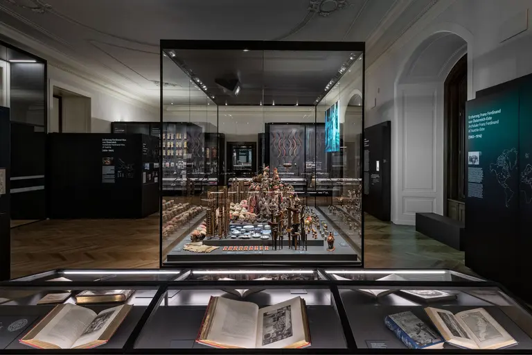 Eine Ausstellung mit einer Glaskabine, die kunstvoll gestaltete Objekte zeigt, umgeben von dunklen Wänden. Im Vordergrund liegen mehrere aufgeschlagene Bücher, die historische Inhalte zeigen. Das Licht betont die Details der Objekte innerhalb der Vitrine.