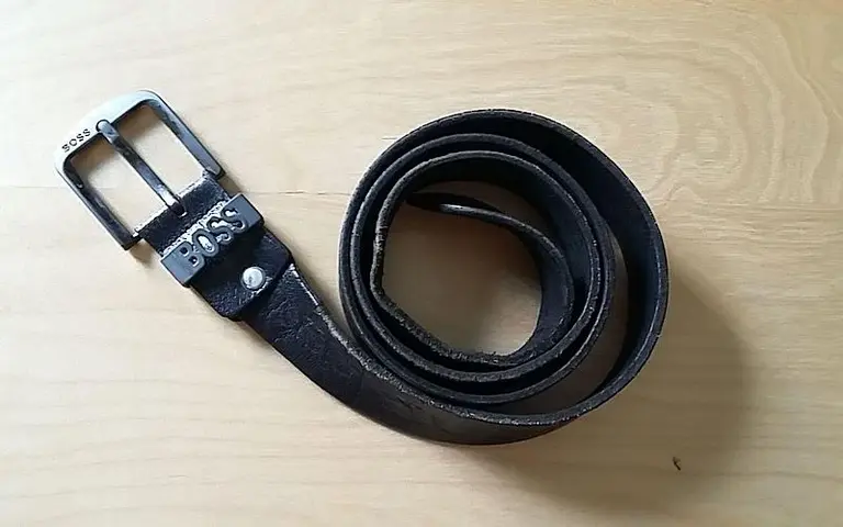Ein schwarzer Ledergürtel liegt auf einer Holzoberfläche. Die Gürtelschnalle ist aus Metall und trägt das Logo 