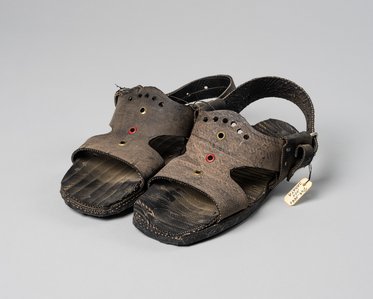 Ein Paar braune Sandalen mit rot und schwarz akzentuierten Lochmustern und einer verstellbaren Ferse. Die Sohle wirkt robust und leicht profiliert. An einer Sandale hängt ein kleines Etikett.