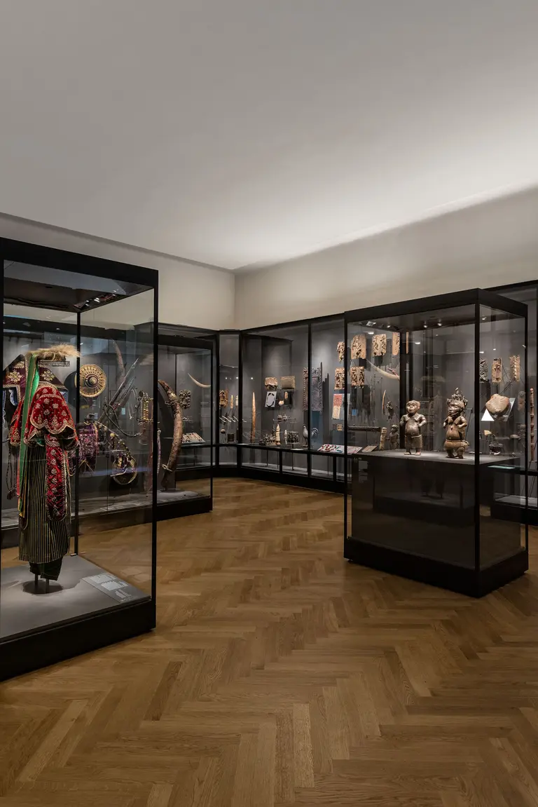 Ein Innenraum eines Museums mit Glasvitrinen, die verschiedene Kunstobjekte und historische Artefakte ausstellen. Der Boden ist aus Holz und die Wände sind neutral bemalt, was die Exponate betont. Die Beleuchtung ist weich und hebt die Details der Ausstellung hervor.