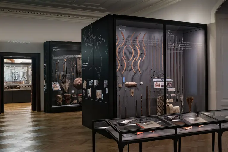 Das Bild zeigt ein Museum mit einer dunklen Vitrine, die verschiedene kulturelle Artefakte und Werkzeuge ausstellt. An den Wänden sind Beschreibungen und Karten angebracht. Der Boden ist aus Holz und die Ausstellungsfläche ist hell beleuchtet.