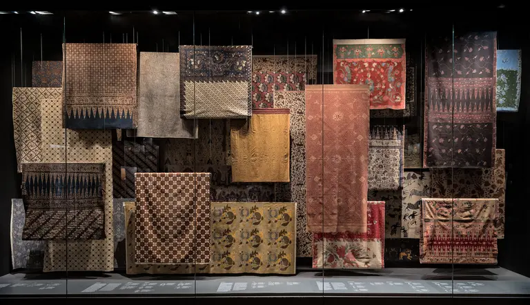 Eine Anordnung von verschiedenen Textilien hängt an einer Wand. Die Stoffe zeigen eine Vielzahl von Mustern und Farben, darunter geometrische Designs und florale Motive. Sie sind in unterschiedlichen Größen und Texturen präsentiert, was einen Eindruck von Vielfalt und Kunstfertigkeit vermittelt.