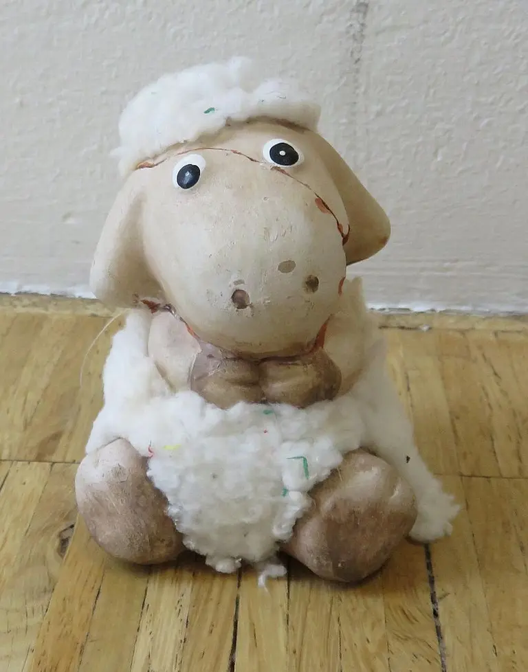Ein niedliches, sitsames Schaf aus Keramik mit einem bemalten Gesicht und einer weichen, wolligen Fellstruktur. Das Schaf hat große, runde Augen und sitzt auf einem hölzernen Boden. Es strahlt eine freundliche und kindliche Ausstrahlung aus.