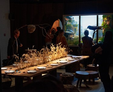 Eine künstlerische Installation zeigt einen langen Tisch mit leeren Tellern und dekorativen Pflanzen. Im Hintergrund sind Menschen zu sehen, die sich in einem Raum mit großen Fenstern aufhalten, die eine malerische Landschaft zeigen. Die Atmosphäre wirkt einladend und kreativ.