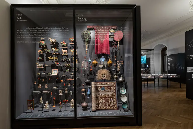 Eine Vitrine zeigt eine Sammlung von Kunst- und Alltagsgegenständen aus Delhi und Bombay. Die Objekte umfassen Figuren, Töpferwaren und kunstvoll gestaltete Behälter, die die kulturelle Vielfalt beider Städte widerspiegeln. Texttafeln informieren über die Herkunft und Bedeutung der Ausstellungsstücke.