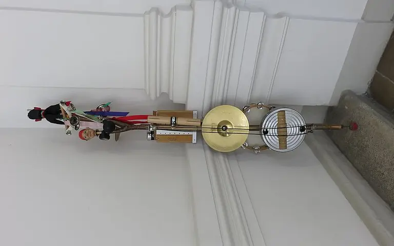 Eine bunte Kunstfigur steht auf einem Sockel, begleitet von verschiedenen Musikinstrumenten. Der obere Teil zeigt eine menschliche Darstellung, während darunter Schlaginstrumente zu sehen sind. Die gesamte Komposition strahlt Kreativität und kulturelle Vielfalt aus.