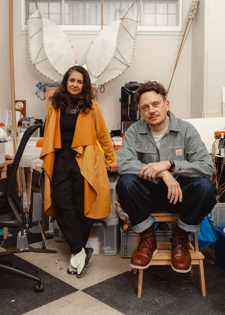 A woman in a yellow coat and a man in a denim jacket sit in an artistic workspace. Behind them are large, abstract wings, and various creative materials are scattered around. The setting suggests a collaborative artistic environment.