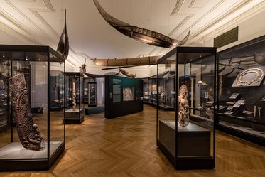 A spacious museum gallery featuring several glass display cases containing various artifacts. The room has wooden flooring and ornate ceilings, with large canoe-like structures suspended overhead. The walls exhibit panels with information about the artifacts, enhancing the educational atmosphere of the space.