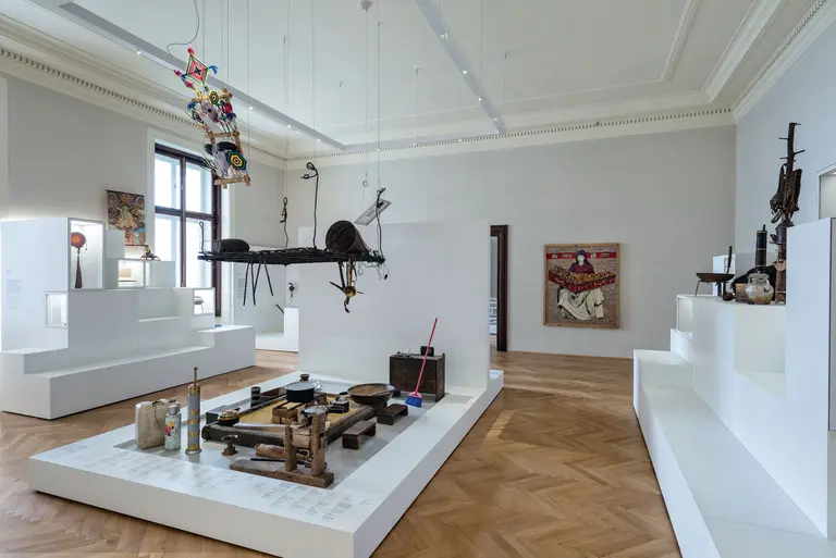 Das Bild zeigt eine moderne Kunstausstellung mit verschiedenen Skulpturen und Objekten, die auf weißen Podesten angeordnet sind. Im Hintergrund ist ein farbenfrohes Kunstwerk an der Wand zu sehen. Der Raum hat große Fenster, die Tageslicht hineinlassen.