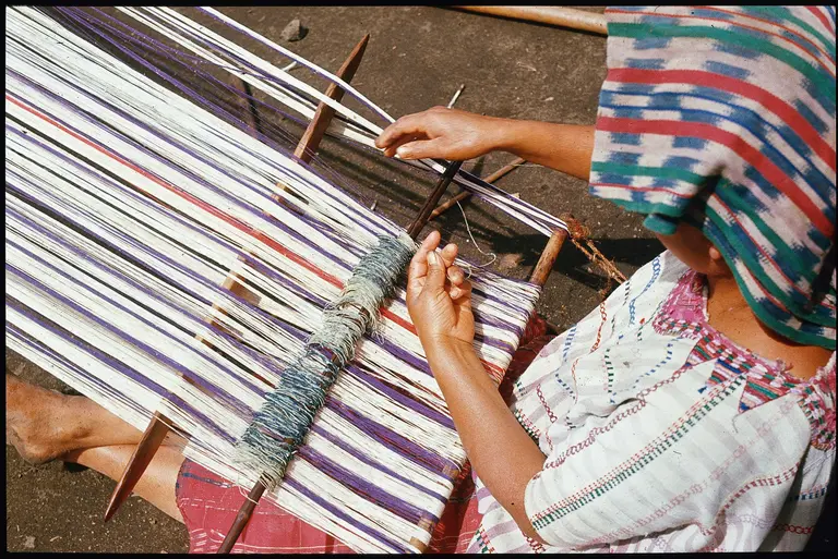 Eine Person arbeitet an einem Webstuhl, auf dem bunte Fäden in lila, blau und weiß angeordnet sind. Die Person trägt ein traditionelles Gewand und eine Kopfbedeckung. Sie hält die Fäden mit einer Hand und nutzt die andere Hand, um das Gewebe zu formen.