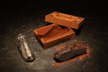 Ein Holzrahmen mit einer Öffnung zeigt eine mit feinem Pulver gefüllte Innenfläche. Neben dem Rahmen liegt ein Stück geschmolzenes Material und ein transparenter Schuh, der leicht diagonal platziert ist. Der Hintergrund ist dunkel und verleiht der Szene eine interessante Stimmung.