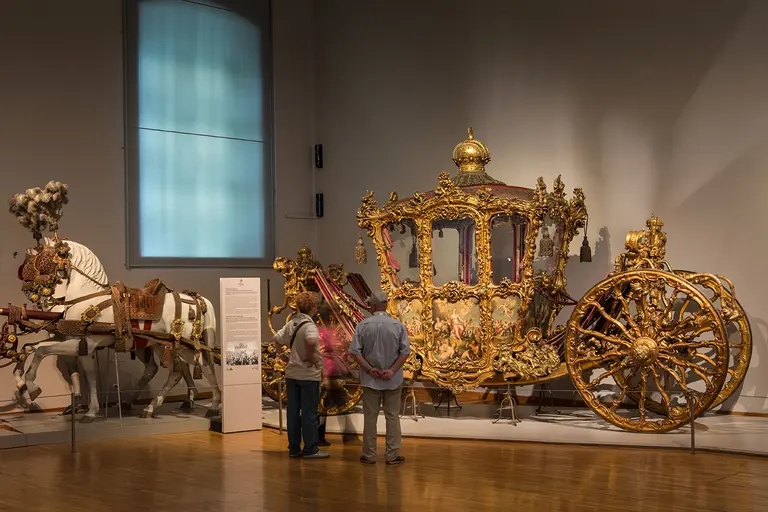 Ein prächtiger, goldverzierter Kutschenwagen steht in einem Museum. Zwei Besucher betrachten ihn, während ein dekoriertes Pferd daneben steht. Die Kutsche ist kunstvoll gestaltet und spiegelt historischen Reichtum wider.