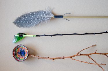 Drei Objekte liegen auf einem neutralen Hintergrund: Ein Pfeil mit einer Feder, ein dünner, bewachsener Zweig mit Dornen und eine buntem Schnur gewickelte Rolle. Jedes Objekt hat unterschiedliche Texturen und Farben.