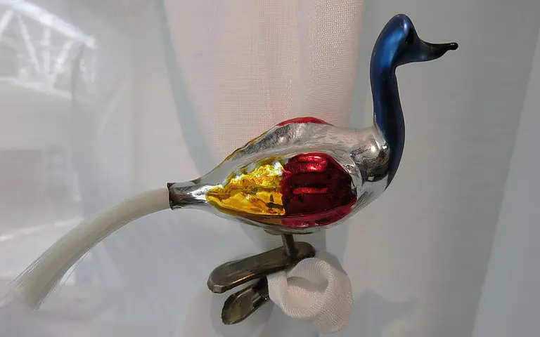 Eine dekorative, metallische Figur in Form eines Vogels. Der Vogel hat einen blauen Kopf, einen glänzenden Körper in Silber, Rot und Gold sowie einen langen, weißen Schwanz. Er sitzt auf einem kleinen Fuß, der an einem Untergrund befestigt ist.