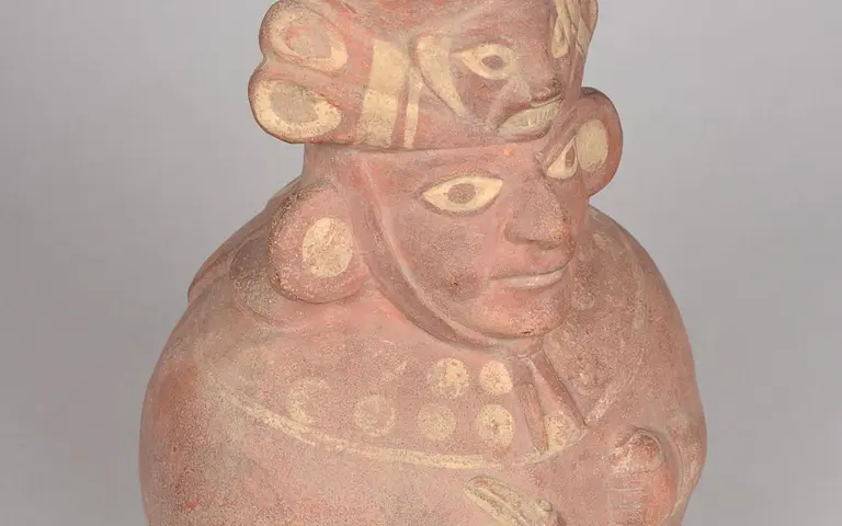 A clay sculpture depicting a human figure with distinct facial features, large eyes, and decorative headgear. The figure is adorned with ear ornaments and intricate patterns, conveying an expression of seriousness or contemplation. The surface has a warm, reddish tone, highlighting its ancient craftsmanship.