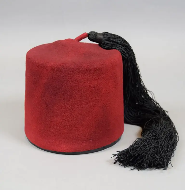 Ein roter Fez mit einer langen schwarzen Quaste. Der Hut hat eine runde Form und eine weiche Oberfläche, die an Filz erinnert. Er wird häufig mit traditioneller Kleidung in bestimmten Kulturen assoziiert.