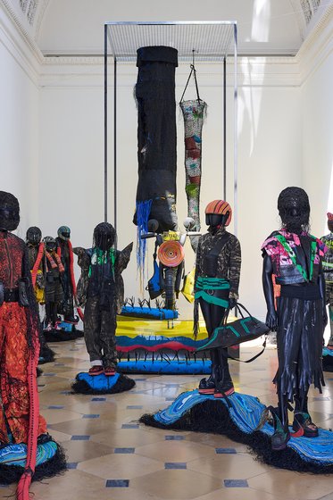 Eine kunstvolle Installation mit zahlreichen menschlichen Figuren aus Textilien in verschiedenen Farben. Einige Figuren tragen leuchtende und verziertete Kostüme, während andere objektspezifische Strukturen in einem hellen Innenraum umgeben. Fokus auf die textile Vielfalt und kreative Gestaltung.