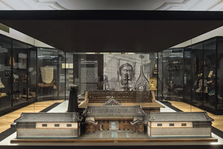 Das Bild zeigt eine historische Ausstellung in einem Museum. Im Vordergrund befindet sich ein detailliertes Modell eines traditionellen Gebäudes, während im Hintergrund eine große informative Tafel mit textuellen Beschreibungen zu sehen ist. Die Vitrinen enthalten weitere historische Objekte.