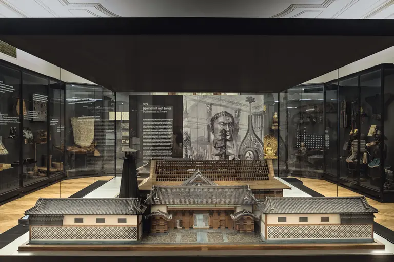 Das Bild zeigt eine historische Ausstellung in einem Museum. Im Vordergrund befindet sich ein detailliertes Modell eines traditionellen Gebäudes, während im Hintergrund eine große informative Tafel mit textuellen Beschreibungen zu sehen ist. Die Vitrinen enthalten weitere historische Objekte.