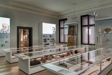 A museum interior featuring several display cases showcasing historical artifacts. Two large screens on the walls display photographs of individuals, and various items are illuminated under glass, highlighting their significance in the exhibition.