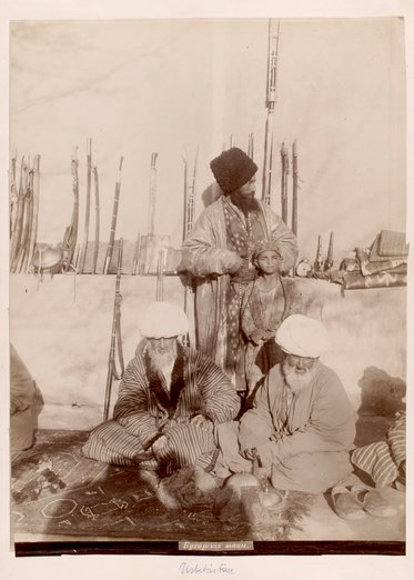 Drei Männer und ein Junge sitzen und stehen in einem Zelt. Die Männer tragen traditionelle Kleidung und haben große Kopfbedeckungen. Im Hintergrund sind verschiedene Gegenstände, darunter Gewehre und Teppiche, sichtbar.
