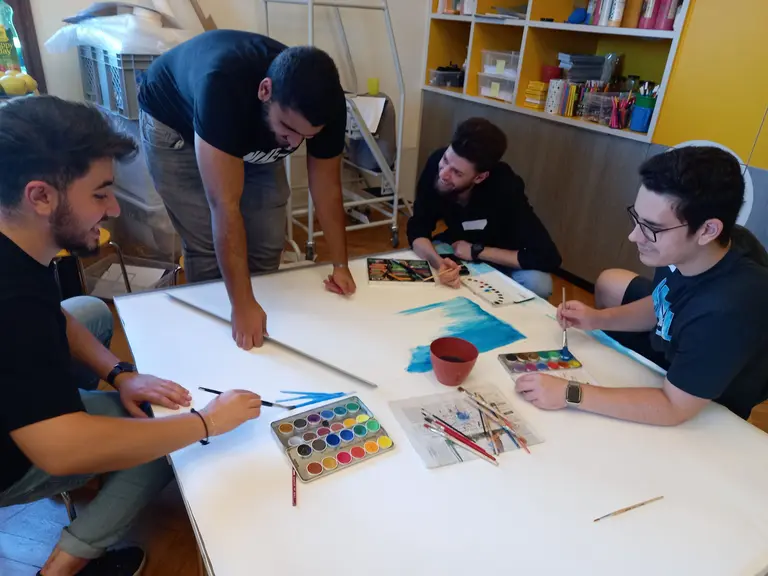 Vier junge Männer sitzen um einen großen Tisch, auf dem Malutensilien liegen. Sie arbeiten gemeinsam an einem Kunstprojekt, das Aquarellfarben und Pinsel beinhaltet. Auf dem Tisch sind kupferne Töpfe und Skizzen, während einer der Männer mit einem Pinsel malt.