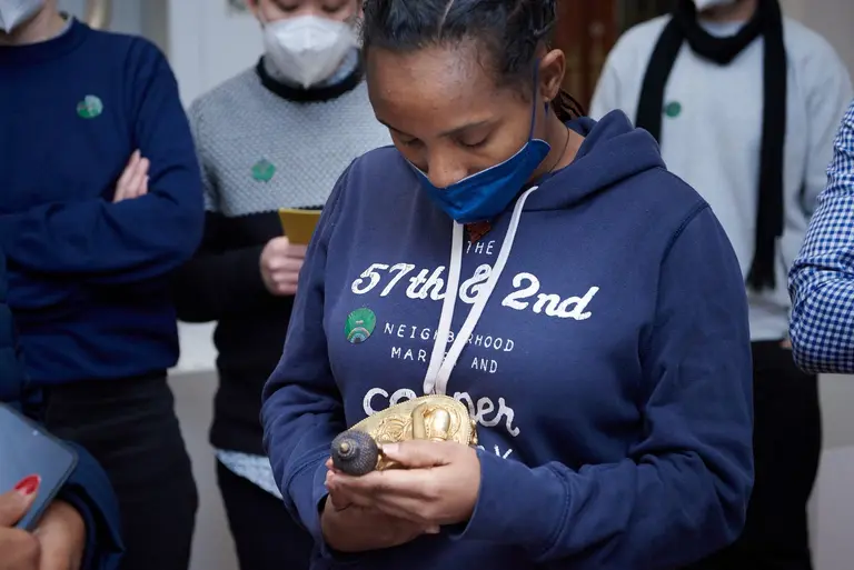 Eine Person mit einem blauen Mundschutz hält eine kleine goldene Statue in beiden Händen. Sie schaut aufmerksam auf das Objekt. Um sie herum stehen weitere Personen in neutraler Kleidung, die ebenfalls interessiert wirken.