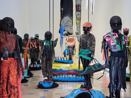Eine Kunstausstellung mit mehreren Statuen, die menschliche Figuren darstellen. Sie sind mit kunstvoll gestalteter Kleidung und langen, haarähnlichen Materialien bedeckt. Im Hintergrund sind farbenfrohe Skulpturen und textile Elemente zu sehen, die die künstlerische Atmosphäre der Installation unterstreichen.