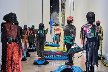 Eine Kunstausstellung mit mehreren Statuen, die menschliche Figuren darstellen. Sie sind mit kunstvoll gestalteter Kleidung und langen, haarähnlichen Materialien bedeckt. Im Hintergrund sind farbenfrohe Skulpturen und textile Elemente zu sehen, die die künstlerische Atmosphäre der Installation unterstreichen.
